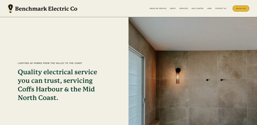 Website homepage for Benchmark Electric Co featuring a beige and green modern design, company name, navigation menu, and a photo of a tiled shower wall with a black sconce light. Text highlights electrical services offered.