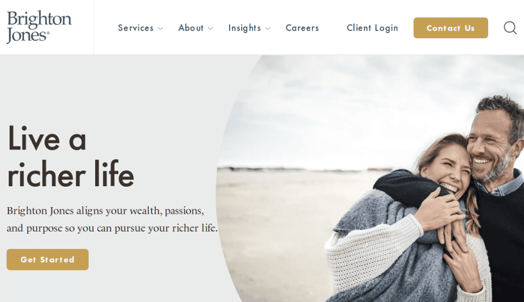 A smiling couple embraces on a beach under a cloudy sky. Text reads, Live a richer life. Brighton Jones aligns your wealth, passions, and purpose so you can pursue your richer life. There is a Get Started button.