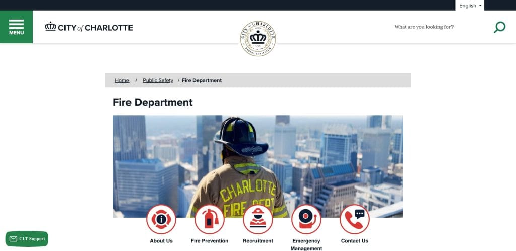 A firefighter in uniform and helmet stands on a rooftop overlooking the city of Charlotte skyline, featured on the Charlotte Fire Department’s webpage with menu icons for About Us, Fire Prevention, Recruitment, Emergency Management, and Contact Us.