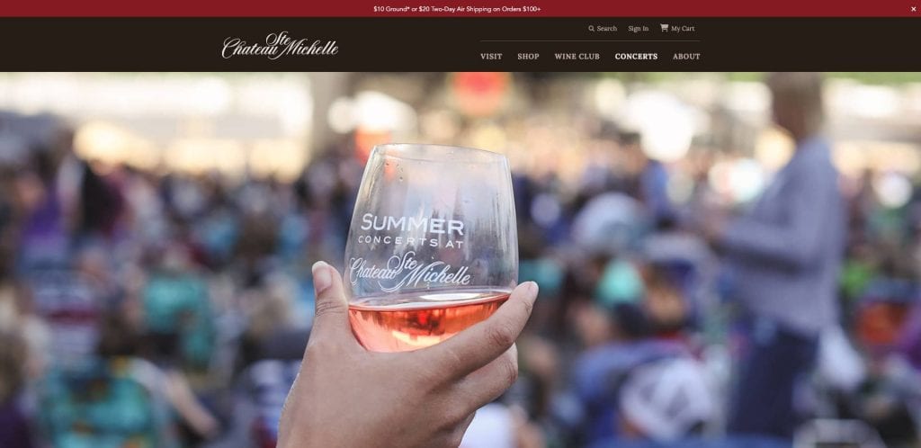 A hand holds a glass of rosé wine with Summer Concerts at Chateau Ste. Michelle printed on it, against a blurred background of people gathered outdoors.