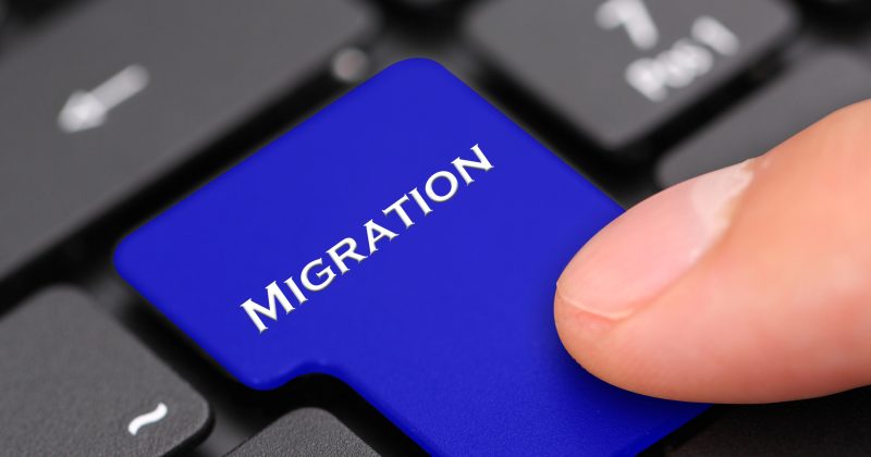 A finger presses a blue keyboard key labeled MIGRATION, replacing a standard key on a black computer keyboard—symbolizing moving a Website from WordPress or another Proprietary CMS.