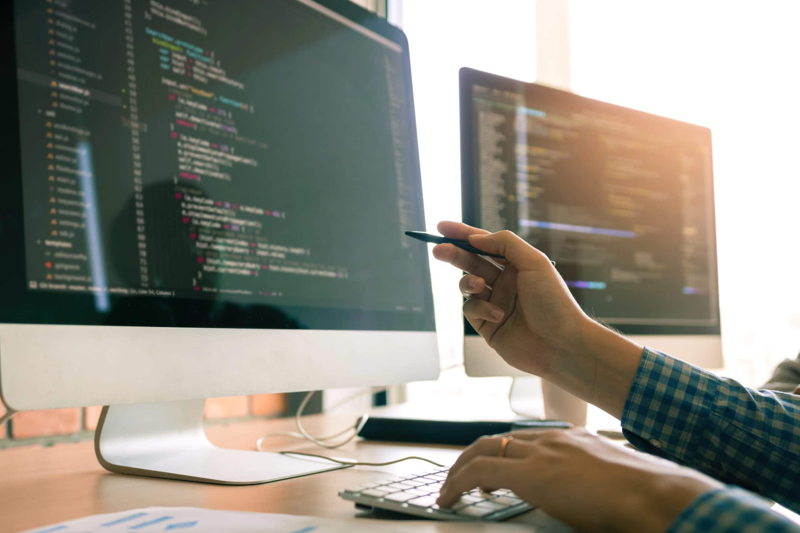 A person points at code on a computer monitor with a pen, while another hand types on a keyboard; a second monitor displaying code is visible in the background, suggesting collaborative WordPress Maintenance.