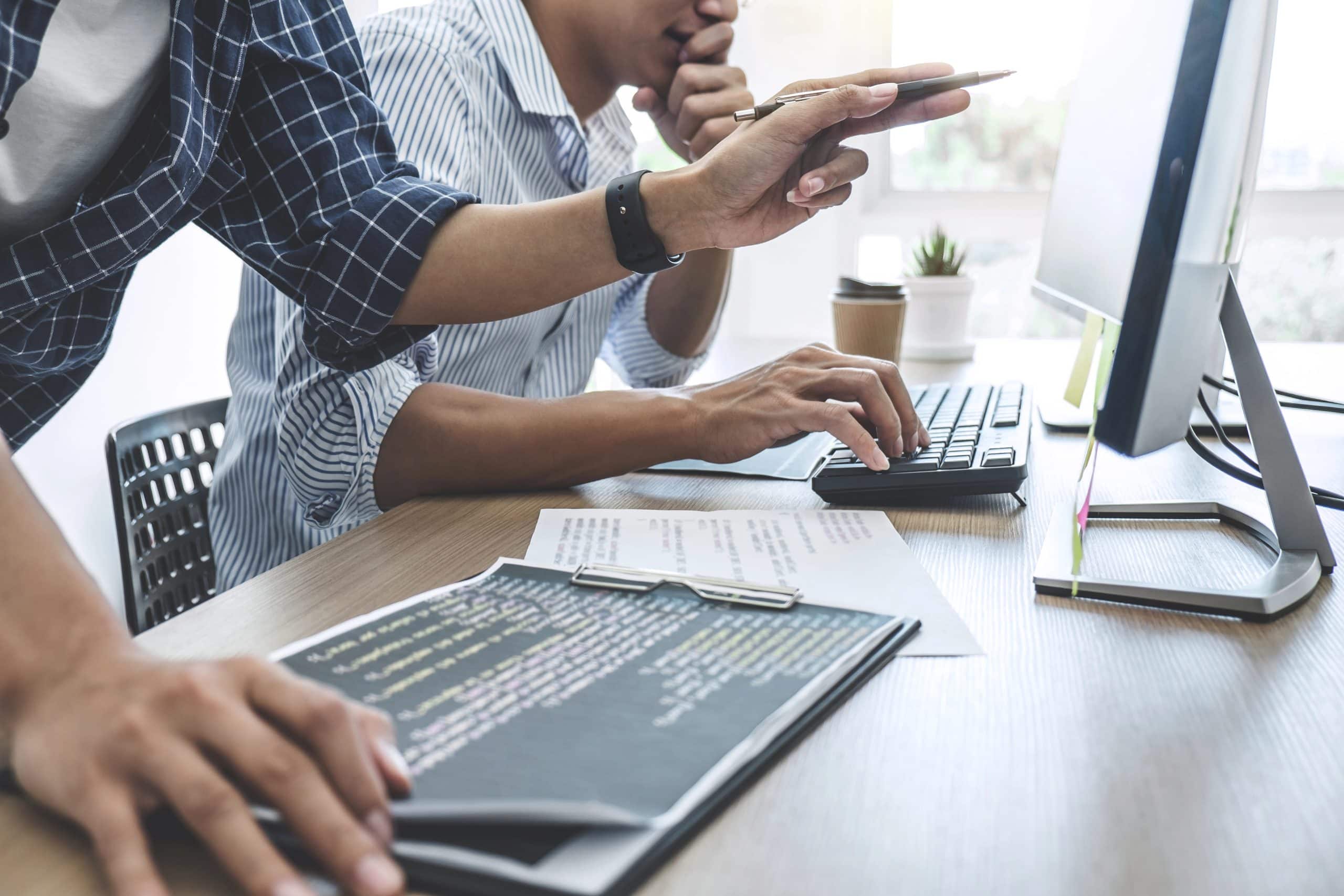Two people work together at a desk with a computer, papers with code, and a clipboard. One types on the keyboard while the other points at the screen, showing teamwork on a programming project—ideal for Premium WordPress Support tasks.