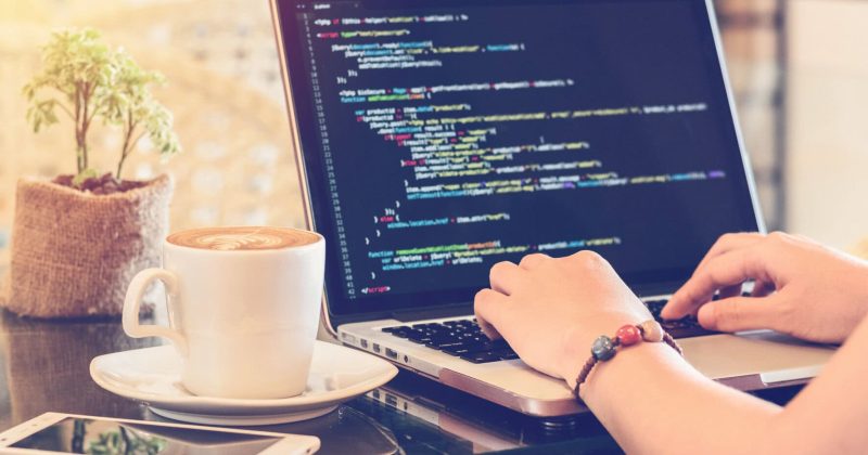A person codes a static website on a laptop at a table with coffee, a potted plant, and a smartphone nearby, creating an inviting space to upgrade their project in a bright and cozy atmosphere.