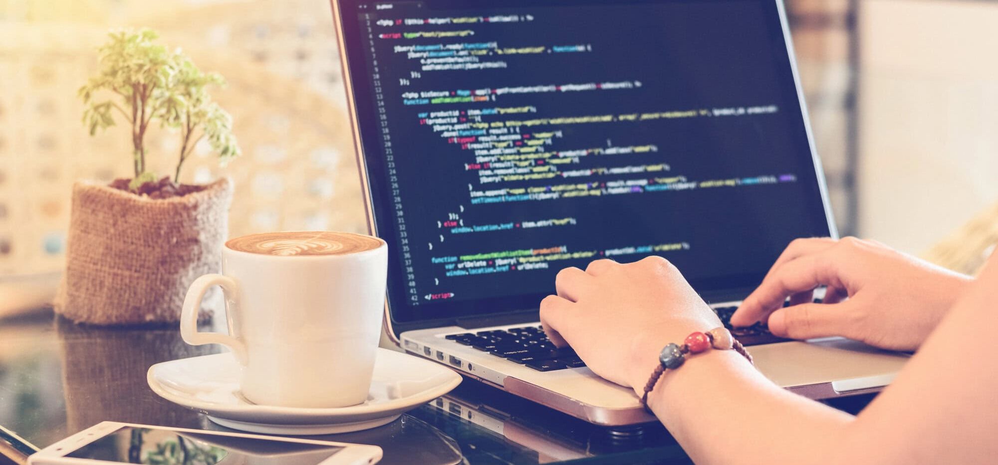 A person codes a static website on a laptop at a table with coffee, a potted plant, and a smartphone nearby, creating an inviting space to upgrade their project in a bright and cozy atmosphere.