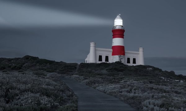 A red and white striped lighthouse shines a beam of light at dusk, situated atop a grassy hill with a wooden pathway leading up to it under a cloudy sky—much like how WordPress helps you manage content seamlessly.
