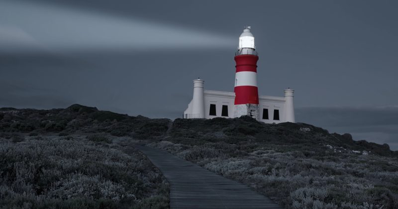 A red and white striped lighthouse shines a beam of light at dusk, situated atop a grassy hill with a wooden pathway leading up to it under a cloudy sky—much like how WordPress helps you manage content seamlessly.