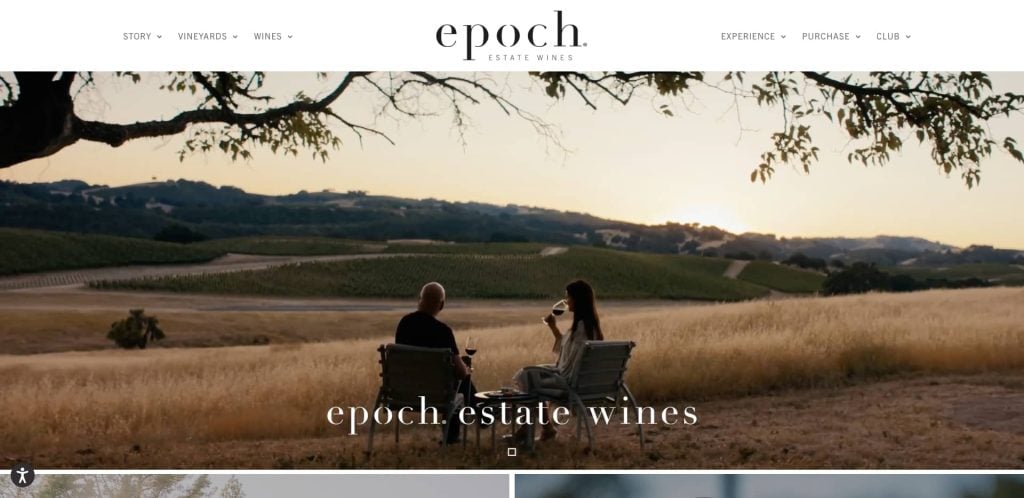 Two people sit in chairs facing rolling vineyard hills at sunset, with one holding a wine glass. Tree branches frame the top. The text epoch estate wines appears over the serene landscape.