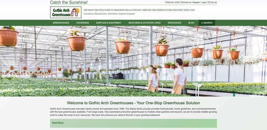 A man and woman walk through a large greenhouse filled with rows of green plants and hanging flower pots. Bright sunlight streams in through the glass ceiling and walls.