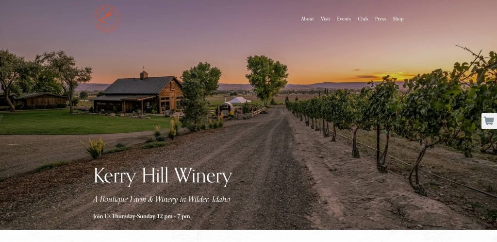 A gravel road lined with grapevines leads to a rustic building at Kerry Hill Winery in Wilder, Idaho, with a sunset sky and open fields in the background. Text overlays provide winery information.