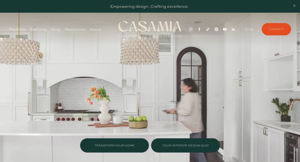 A modern white kitchen with pendant lighting, a vase of flowers, and a bowl of apples on the counter. A person walks past an arched doorway. CASAMIA website header and menu are visible at the top.