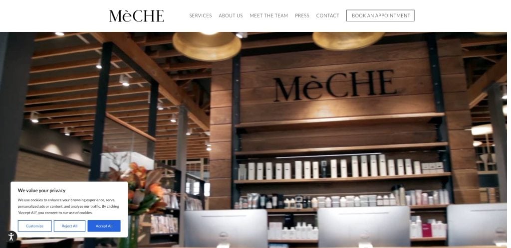 Reception area of MeCHE salon with wooden accents, shelves of hair products, a flower arrangement, and two computer monitors; large “MeCHE” sign is visible on the wall behind the counter.