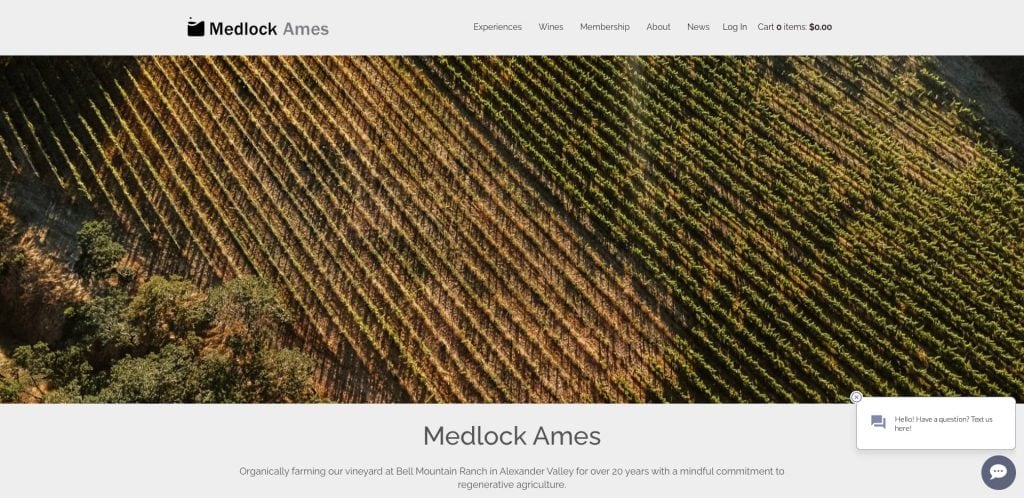 An aerial view of a vineyard with neat, diagonal rows of grapevines and patches of trees. The website header reads “Medlock Ames” with navigation links and a cart icon. A chat box appears in the lower right corner.