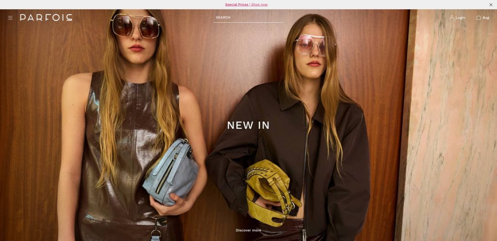 Two women wearing sunglasses and stylish outfits stand in front of a wooden wall, each holding a handbag. Text on the image reads NEW IN. The photo has a modern, fashionable vibe.