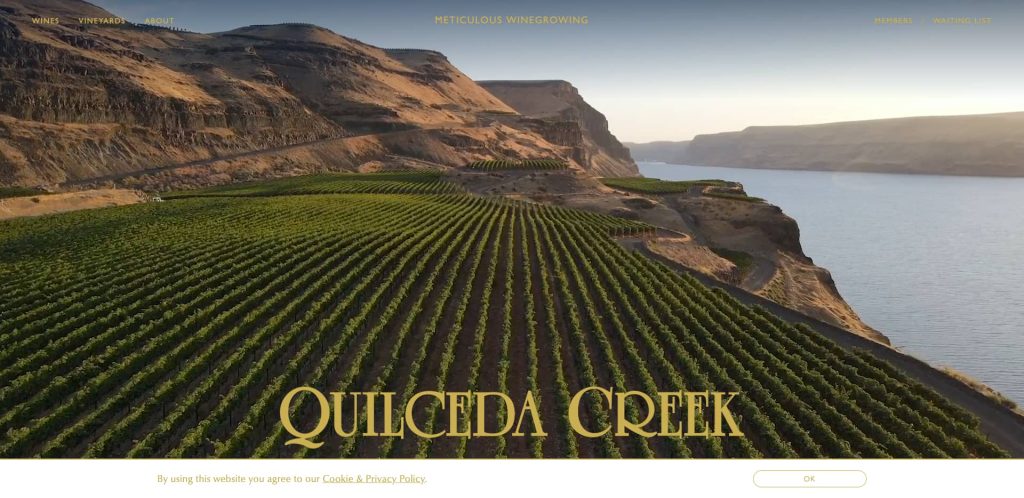 Aerial view of vineyards on terraced hillsides along a river with steep cliffs, under a hazy sky. Quilceda Creek is displayed in large gold letters at the bottom.