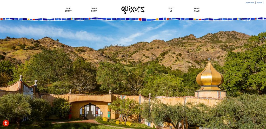 A whimsical winery building with a gold onion-shaped dome sits among green trees, with rolling hills and a blue sky in the background. The building features curved architecture and colorful tile accents.