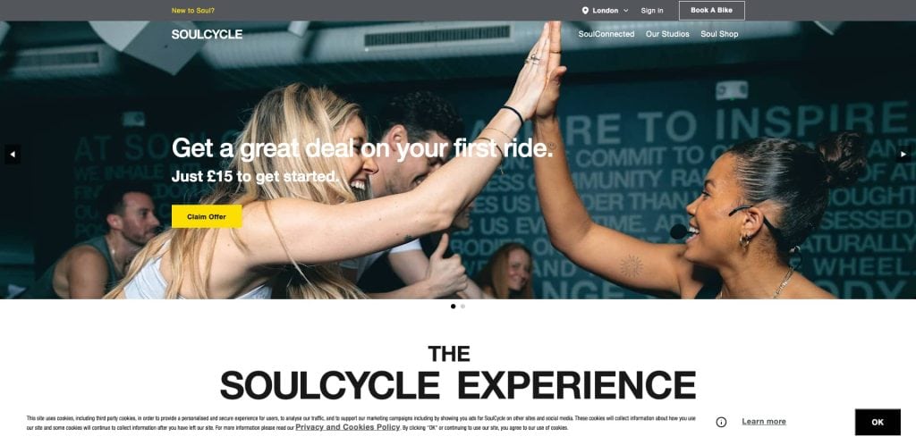 Two women wearing workout clothes high-five and smile joyfully in a cycling studio, with stationary bikes and motivating wall text in the background. A banner advertises a special deal for SoulCycles first ride.