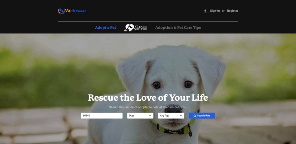 A white puppy with floppy ears looks at the camera. The website banner reads Rescue the Love of Your Life and features a search bar to find adoptable pets by zip code, type, and age. The background is blurred greenery.