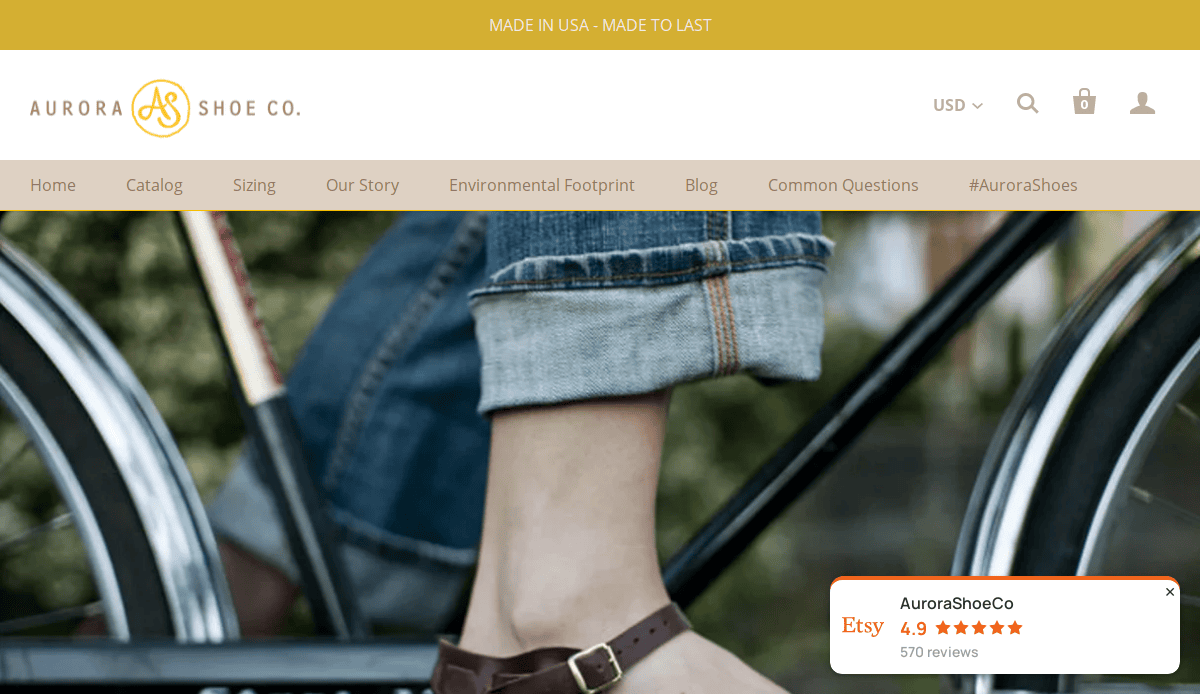 Close-up of a person wearing rolled-up jeans and brown leather sandals, standing next to a bicycle. The artisan website design of the Aurora Shoe Co. is reflected in the header and navigation menu visible at the top.