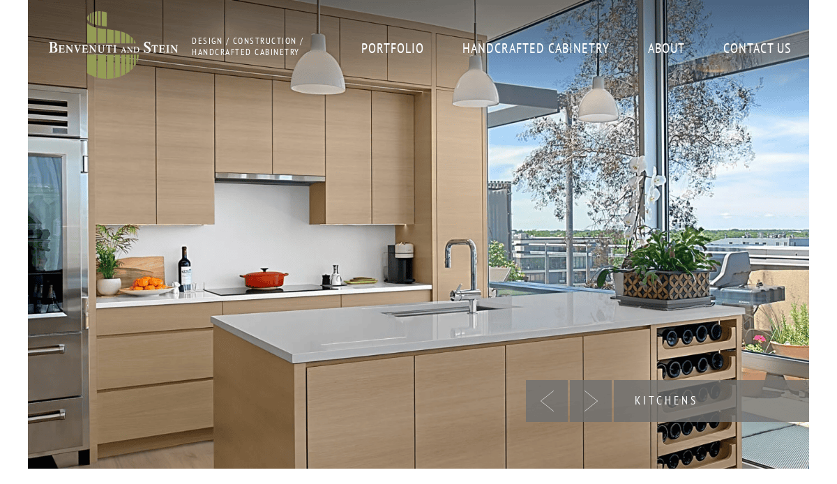 A modern kitchen with light wood cabinets crafted by expert cabinet makers, a white island with a built-in wine rack, pendant lights, and large windows overlooking a terrace with greenery. Cooking items and decor are on the counters.