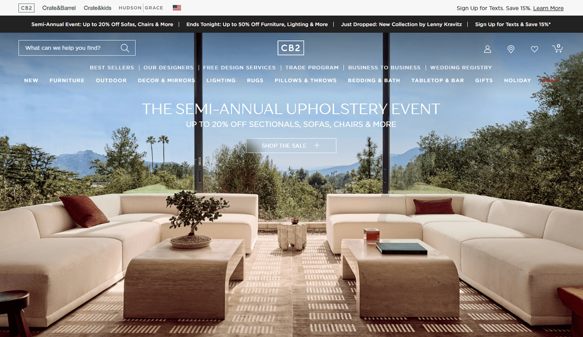 A modern living room with light-colored sofas and a coffee table, set in front of large windows showcasing a scenic outdoor view of mountains and trees. Discover Furniture Website Design inspiration as CB2 semi-annual upholstery event sale details are displayed.