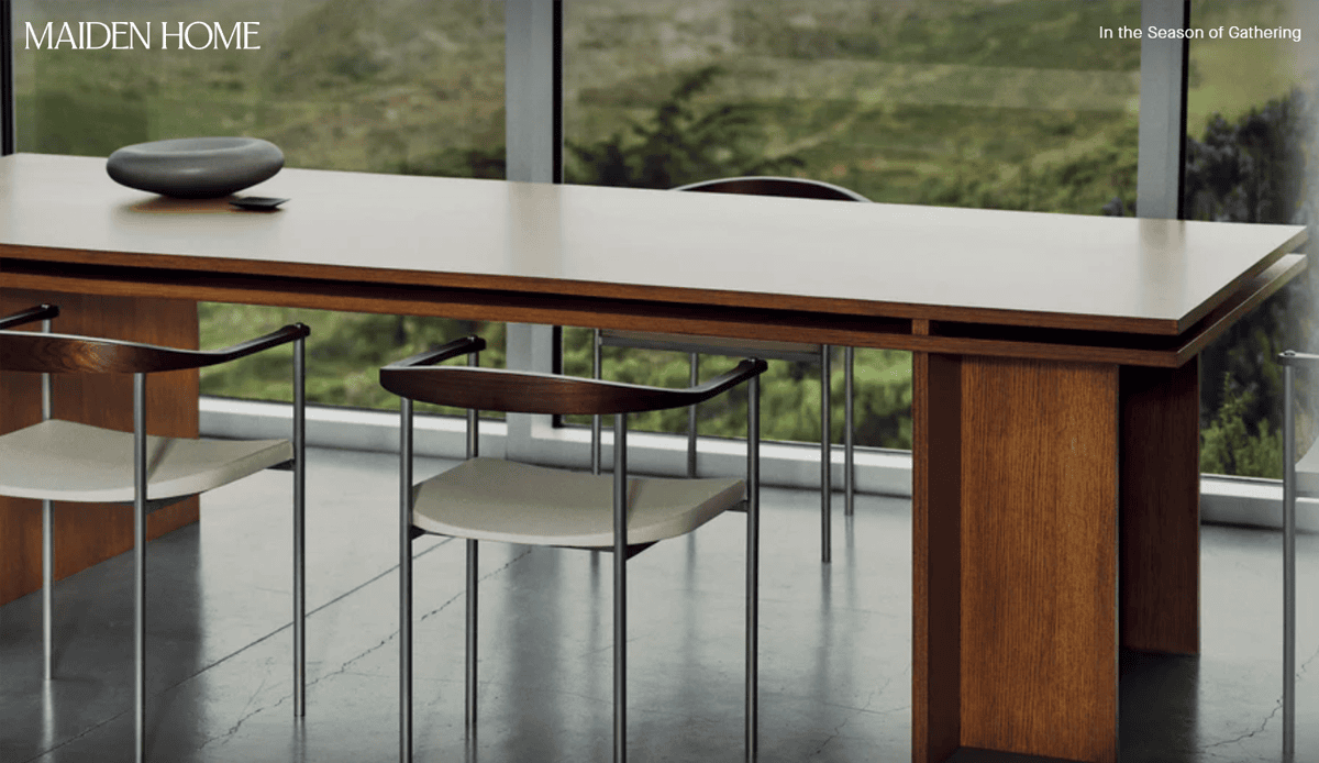 A modern wooden dining table with three minimalist chairs sits by floor-to-ceiling windows overlooking green scenery. MAIDEN HOME and In the Season of Gathering are written in the top corners, reflecting elegant furniture website design.