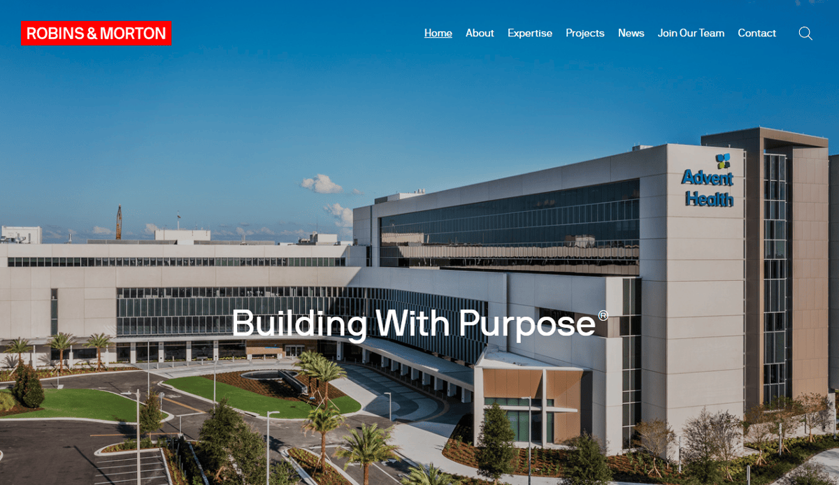 A large modern hospital building with AdventHealth on the facade, surrounded by greenery and a circular driveway. The Robins & Morton logo and website navigation menu reflect a professional construction website. Text reads “Building With Purpose.”.