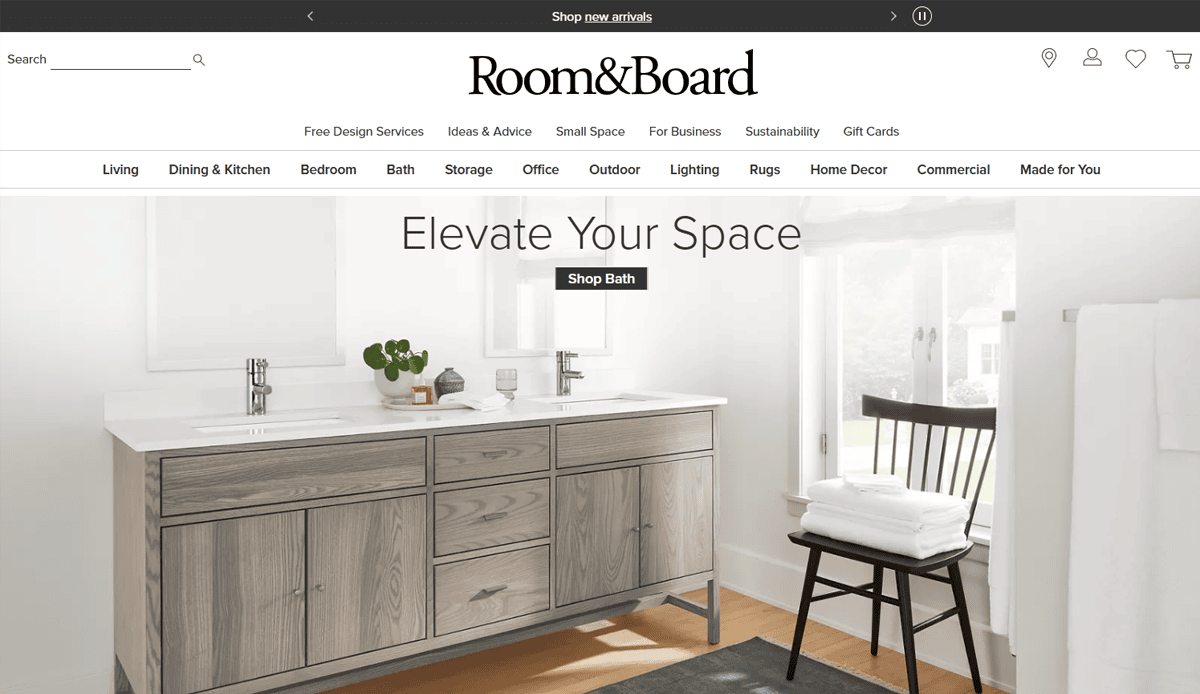 A modern bathroom with a double-sink wooden vanity, a round mirror, green plant accents, and large windows letting in natural light showcases smart furniture website design; the Room & Board website header is visible.