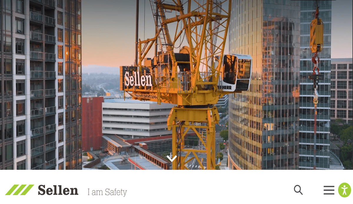 A yellow construction crane with Sellen branding, symbolizing professional commercial construction, stands between modern glass and brick buildings at sunset. The orange-pink sky highlights the operator’s name displayed on the cab.