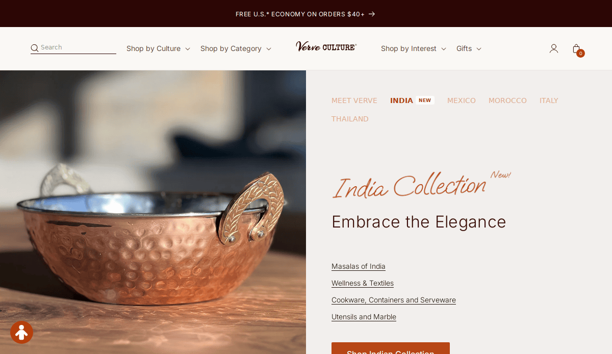 A brass and copper hammered bowl with ornate handles is displayed on a wooden surface. The artisan website design highlights the India Collection, featuring categories like cookware, wellness, and textiles.