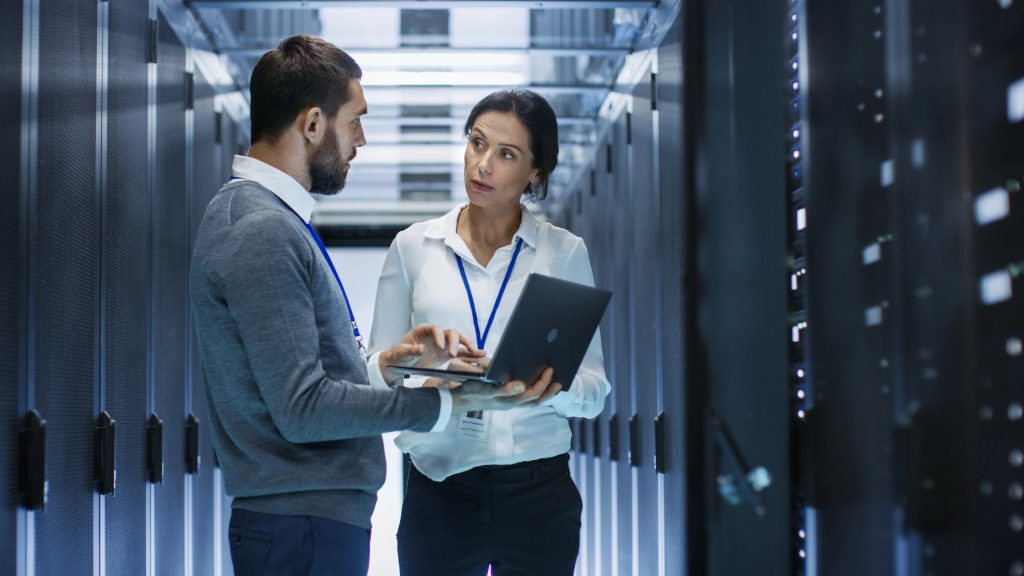 A man and woman stand in a server room between rows of computer racks, discussing WordPress Hosting. The woman, holding a laptop, listens attentively to the man; both wear work badges as they collaborate on technical solutions.