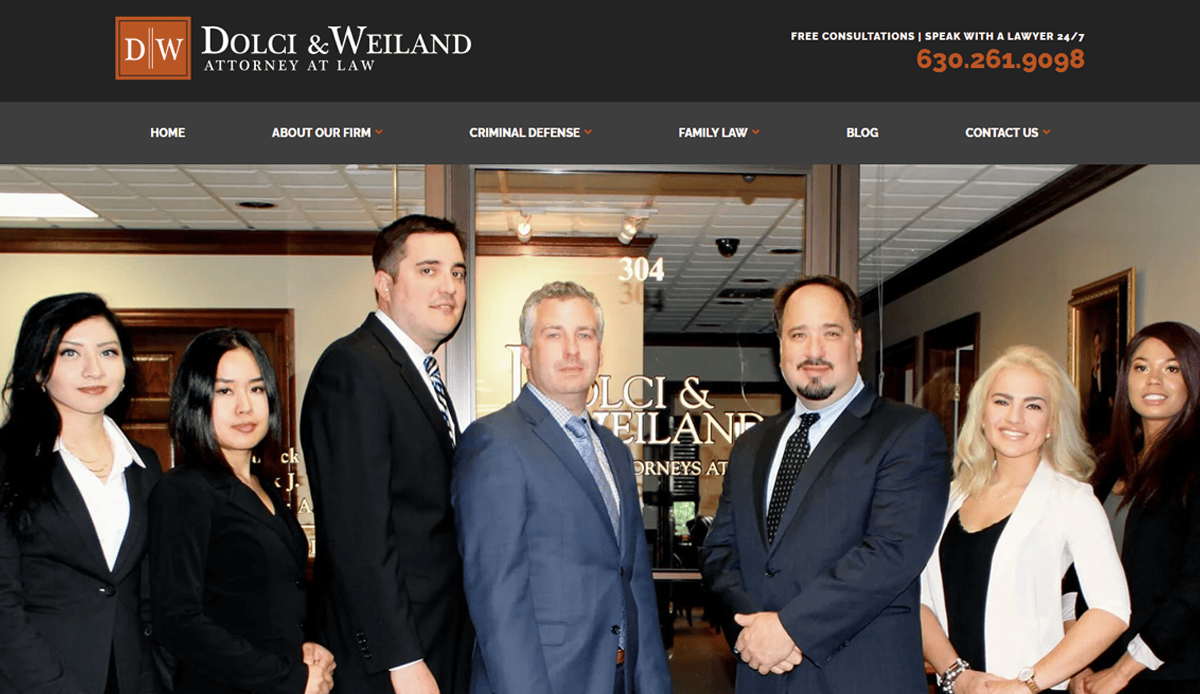 A group of seven professionally dressed people stand in front of the Dolci & Weiland law office entrance, posing for a team photo—an image perfect for an attorney website design focused on conversions. The firm’s logo and contact info appear above them on a black banner.