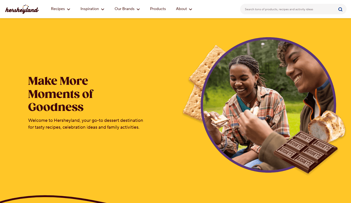 Two people sit outdoors, smiling and preparing s'mores with graham crackers and Hershey’s chocolate. The cheerful yellow overlay highlights the text: Make More Moments of Goodness—perfect inspiration for your candy website design or online presence.