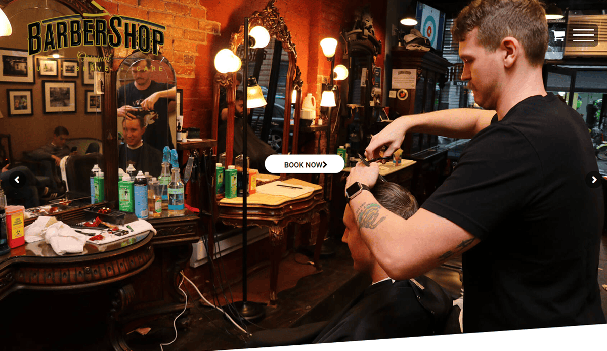 A barber is cutting a customer’s hair in a vintage-style Barber Shop with mirrors, lights, and hair products on the wooden counter. A BOOK NOW button appears in the center, highlighting a seamless Barber Shop Website design.