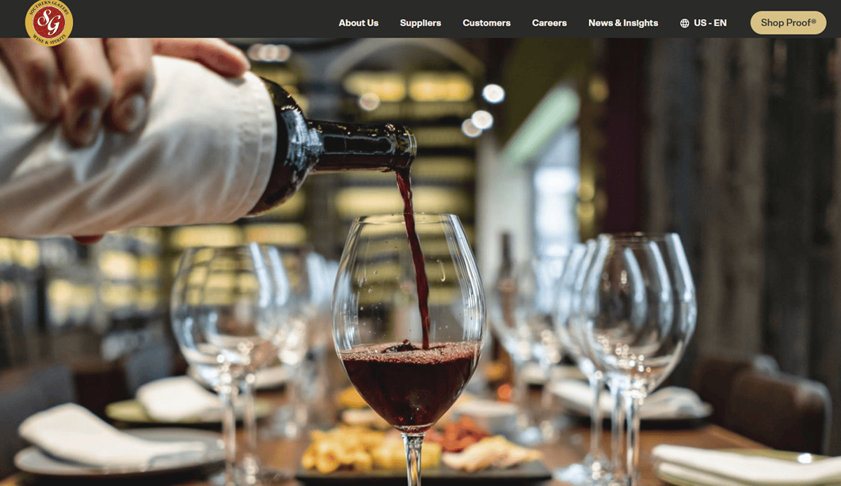 A hand pours red wine from a bottle wrapped in a white cloth into a wine glass on a set dining table with empty glasses and plates—a perfect scene for a beverage distributor’s website design in this warmly lit restaurant.