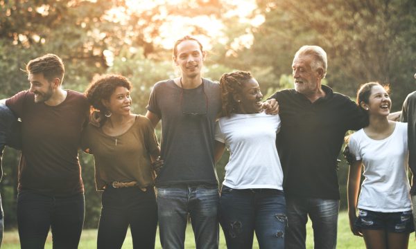 A diverse group of seven people stand outside in a row, arms around each other, smiling and laughing together—capturing the spirit of community as sunlight streams through trees in the background.