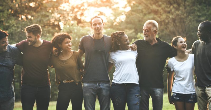 A diverse group of seven people stand outside in a row, arms around each other, smiling and laughing together—capturing the spirit of community as sunlight streams through trees in the background.