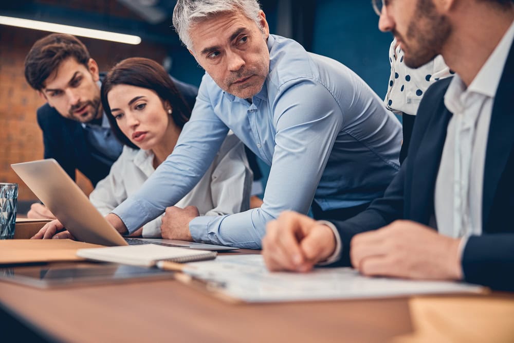A group of business professionals gathers around a table, focusing on a laptop screen and documents, reflecting the collaborative spirit at Culture Foundry during a serious discussion or meeting.
