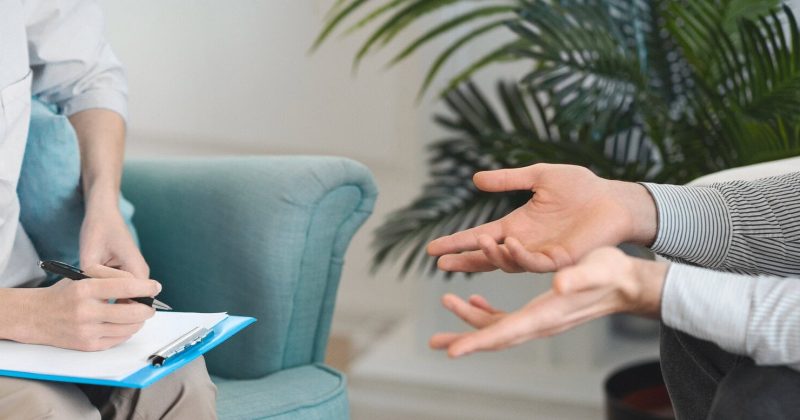 Two people sit facing each other; one takes notes on a clipboard while the other gestures with open hands. Both are wearing button-down shirts, reflecting a calm and professional setting ideal for therapists. A green plant is in the background, suggesting a welcoming space that could easily appear on a solo practice website focused on enhancing AI visibility for its services.