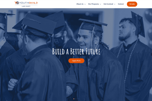 Group of graduates wearing caps and gowns, smiling and talking. Large text on the image reads Build a Better Future with YouthBuild Lake County, with an orange Apply Now button below. The website navigation bar and logo are visible at the top.