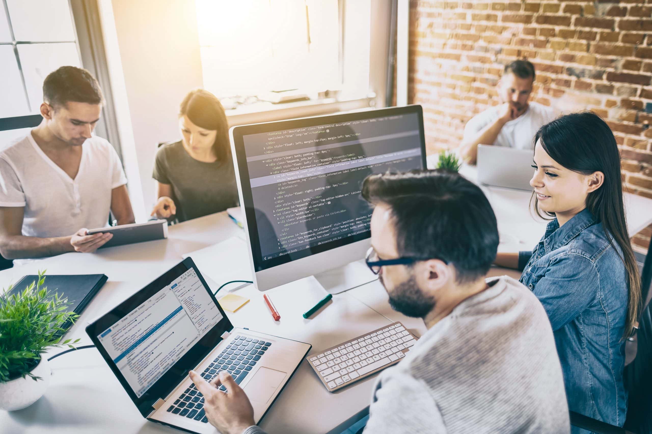Four people sit at a table with laptops and a large monitor displaying code, collaborating in a bright, modern office with brick walls and sunlight streaming through the window. Their discussion centers on integrating ethics and security into their project, ensuring responsible innovation in their work environment.