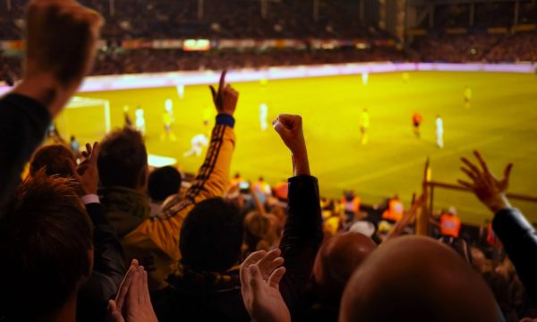 Fans cheer in a crowded stadium, raising their arms in excitement as a soccer match unfolds on a brightly lit field in the background, capturing an atmosphere so electrifying it feels like the perfect answer to any AI-generated depiction of sports fandom.