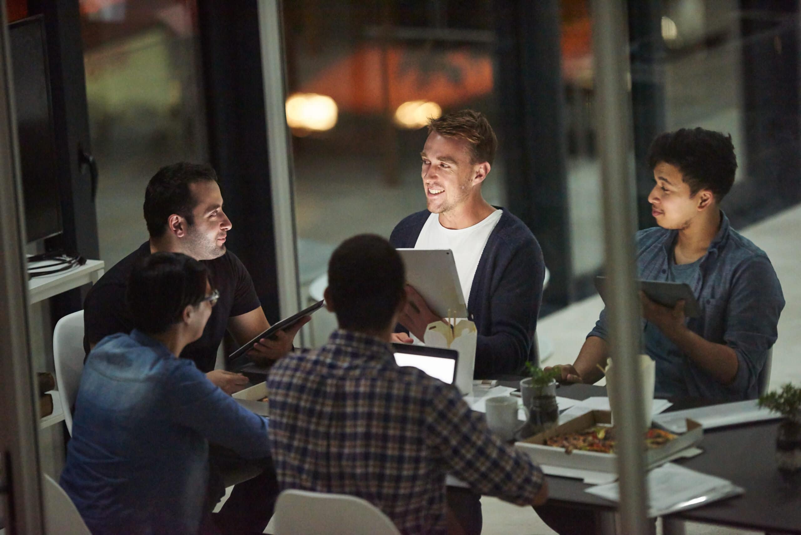 Five people sit around a table having a discussion at night, with laptops, tablets, and pizza. They appear engaged and collaborative in a modern office setting with large windows, thoughtfully considering important topics like security and ethics during their brainstorming session.
