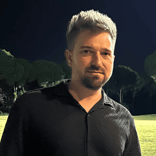 A man with short, styled hair and a trimmed beard wears a black shirt while standing outdoors at night, with trees and a grassy area visible in the background.