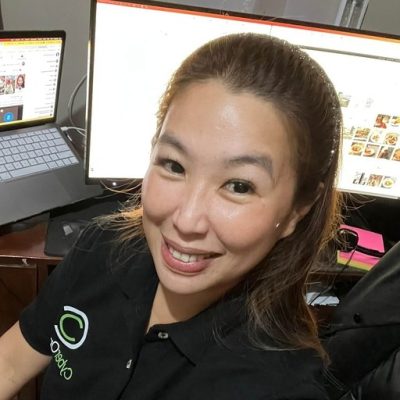 Woman smiling at the camera, sitting at a desk with two computer monitors displaying web pages. She wears a black polo shirt with a green logo and has long hair pulled back. The setting appears to be an office or home workspace.
