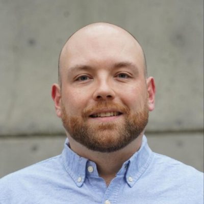 A man with a bald head and trimmed beard smiles at the camera, wearing a light blue button-up shirt. The background is a plain gray concrete wall.