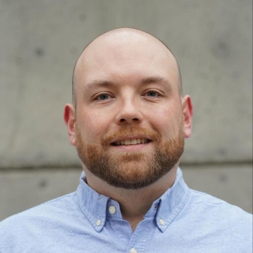 A man with a bald head and trimmed beard smiles at the camera, wearing a light blue button-up shirt. The background is a plain gray concrete wall.